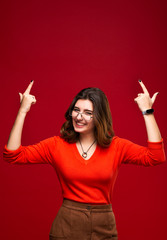 Charismatic smiling girl with glasses shows her fingers up on important information. Place for text. Isolated on red background