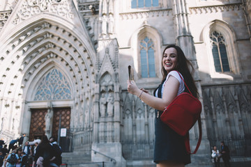 Portrait of cheerful caucasian female traveler excited with exploring old city architecture during vacation journey, smiling beautiful woman 20s tourist standing on square with backpack and map