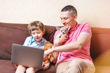 Happy young father sit on couch using laptop relax with preschooler son holding puppy chihuahua have fun together, smiling dad and little boy child enjoy weekend home rest on sofa busy with gadgets