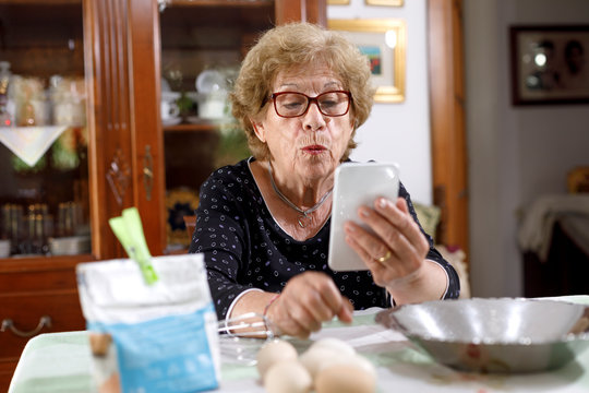 signora anziana con occhiali rossi seduta in una tavola dove stava preparando un dolce fa una videochiamata con il suo smartphone