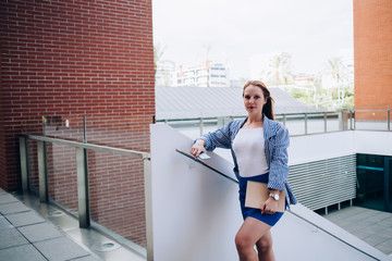 Portrait of redhead caucasian female student walking on stairs in college campus with notepad, young attractive woman 20s looking at camera strolling on urban setting architecture