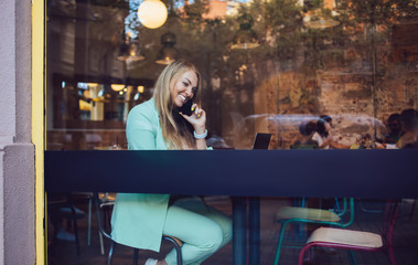 Window view on joyful female freelancer in smart casual wear laughing during positive cellular conversation in cafe interior, cheerful woman using smartphone device for making contact calling