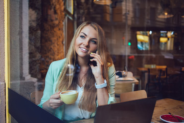 Pretty female freelancer in smart casual wear enjoying coffee during cellular conversation in cafe interior, woman with modern laptop computer using smartphone device for making contact calling