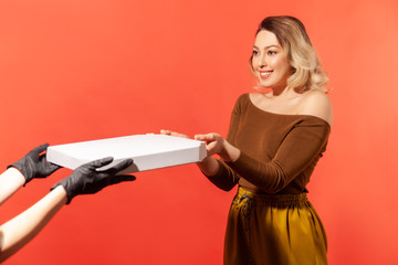 Fototapeta premium Woman taking pizza from courier hands in protective gloves, safe food delivery on quarantine, coronavirus 2019-ncov prevention. Stay home, order online. indoor studio shot isolated on red background