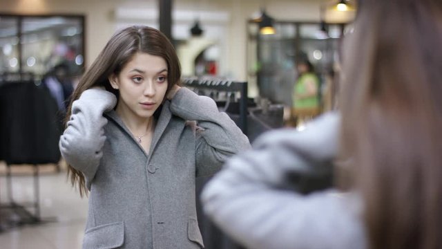 A close up shooting over the shoulder of a young dark haired woman in a clothing boutique looking at herself in the mirror and deciding in buying an overcoat