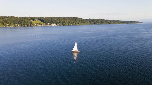 Sideways flight past a small sailing boat on Lake Starnberg. Enthusiastic sailors go sailing in their free time on the mountain lake near Munich. Breathtakingly beautiful aerial view.
