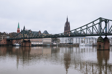 Outdoor cloudy view of Eiserner Steg, historical pedestrian Iron Bridge, and promenade on riverside of Main River with skyline in rainy day in Frankfurt, Germany.