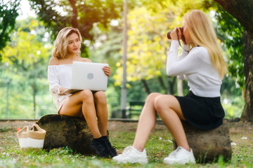 Young caucasian women sitting in natural park, working in a garden with friend and picnic basket and holding laptop in hands. Girl is posing action while her friend shoot a photo, feeling relax.