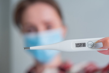 Woman in medical face mask showing digital medical thermometer with high temperature - selective focus, close up. Healthcare, measurement, disease, covid 19, infection, coronavirus concept
