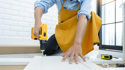 Close up portrait and details of caucasian male worker using electric screwdriver instrument in hand and repairing new wooden desk, home improvement concept.