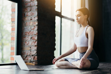 A girl of Asian appearance sits in a lotus position with a laptop and conducts an online yoga training