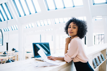Portrait of beautiful female owner of corporation holding financial documentation sitting in office company, confident businesswoman in trendy formal wear spending time for checking accounting report