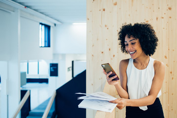 Happy dark skinned female entrepreneur laughing from content in received email from friend via application on mobile phone during break from analyzing paper accounting, smiling woman messaging