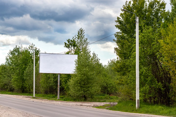 Large blank white billboard on the side of a country road among green trees under a dramatic cloudy sky