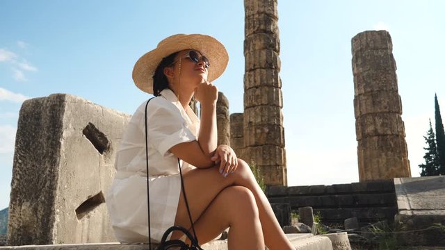 Young woman sitting resting at ruins of temple of Apollo in Delphi, archaeological site by mount Parnassus, Greece, Europe. Large hat, fashion white dress.