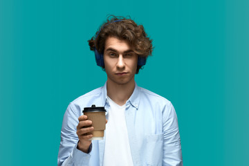 Fototapeta premium Need energy for wake up. Portrait of a young beautiful drowsiness man wearing white t-shirt and blue shirt in headphones with hot drink cup in hand