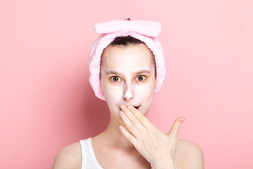 Woman with cosmetic mask on face is surprised to open mouth and cover mouth with hand on pink...