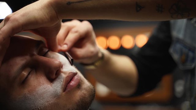 Professional barber shaves customer beard with straight razor. Beard cut with old-fashioned blade at barbershop. Handsome macho man getting his beard shaved in studio. Close-up shot.