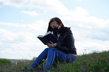 Close up of young woman in black medical mask and casual clothes sitting on hill and reading book on cloudy day. Adult female resting alone in countryside.