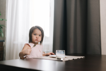 Angry beautiful little two-year-old girl wearing white dress is eating tasty cheesecake with fork for lunch at the table in kitchen room. Glass of milk on the table.