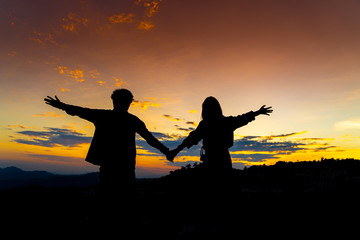 Fototapeta na wymiar Silhouette of Young Asian Couple Holding Hand at Sunset on Mountain with Orange Sky. Enjoying Peaceful Moment Concept. Relaxing or Love Family Relationship Concept.