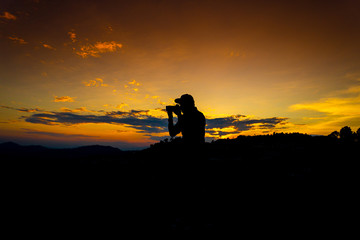 Silhouette Man Standing on Hill with  Camera at the Sunset on Mountain with Orange Sky. Enjoying Peaceful Moment Concept.