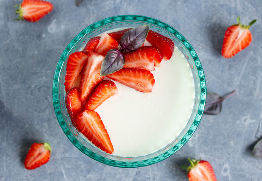 Top view on panna cotta in a turquoise glass bowl with slices of fresh strawberries, basil leaves on a concrete surface