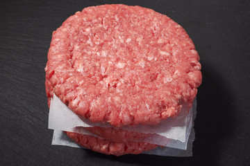 Stack of fresh uncooked beef burgers separated by cooking paper on a black stone plate.