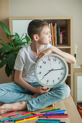 Teenager boy in a white T-shirt sits on a table in a children's room with legs crossed, leaned on a large silver watch. Looks out the window.