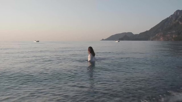 Vacation travel bikini woman walking on beach into water. Beautiful young woman in a white wet blouse in sea water. Happy woman tourist smiling enjoys the warm waves at sunset