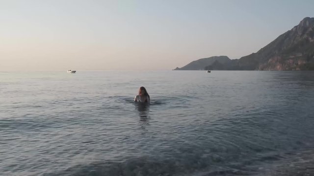 Girl in a white swimsuit comes out of the sea, woman in white wet blouse enjoys a vacation at sea mountain view at sunset