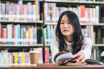Asian young student in white casual suit reading  the book with coffee cup in library of university or colleage.Sitting and reading on a wooden table and bookcase in the background.Back to school