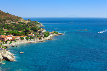 Fototapeta na wymiar Sea shore with beach and rocks and rocky slope of the Island of Elba in Italy. Many people on the beach sunbathing. Blue sea with aerial view. Dwellings of a small village.