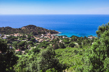Seashore coastline with beach and rocks and rocky slope of the Island of Elba in Italy. Many people on the beach sunbathing. Blue sea with aerial view. Dwellings of a small village.