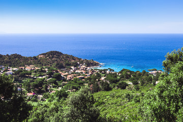 Fototapeta na wymiar Seashore coastline with beach and rocks and rocky slope of the Island of Elba in Italy. Many people on the beach sunbathing. Blue sea with aerial view. Dwellings of a small village.