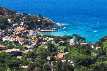 Fototapeta premium Seashore coastline with beach and little village in the Island of Elba in Italy. Many people on the beach sunbathing. Blue sea with aerial view. Houses in the middle of a natural landscape.