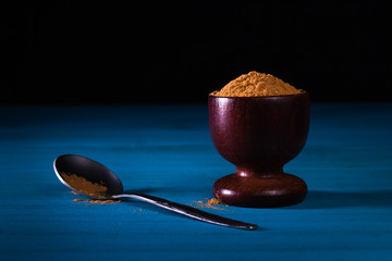 Ground cinnamon powder in wooden cup with small metal spoon on blue wooden table with dark background. Close up.