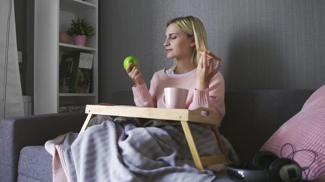 Young woman sits on couch and makes a choice what to eat toast with chocolate or apple