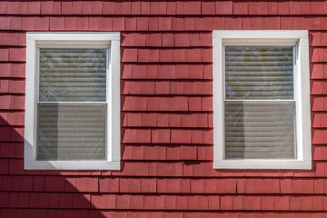 Two white sash windows with shade on a house wall covered with red shake and shingle siding made of wood