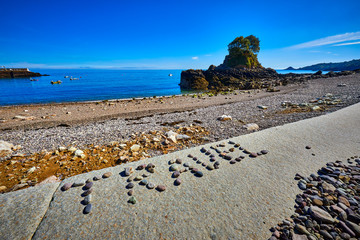 Image of a pebble beach in the early morning sunshine with a small island and harbour in the background with the words TRAVEL written in pebbles in the forground. Selective Focus