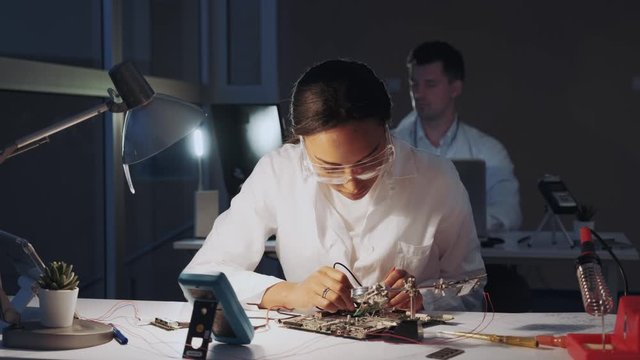African american electronics engineers checking electronic board with multimeter tester and other electronic devises in lab. There is a fellow worker in the background.