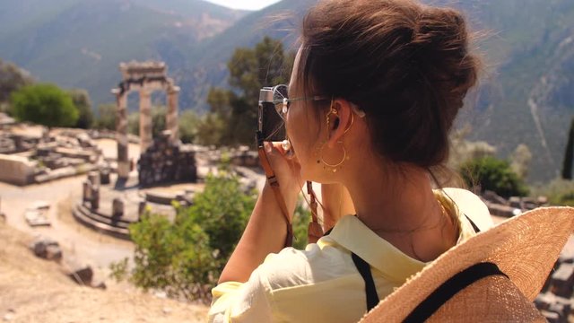 Young woman tourist taking pictures at temple of Athena Pronaia in archaeological site of Delphi, Voiotia, Greece. Fashion elegant yellow dress, large hat, vintage camera.