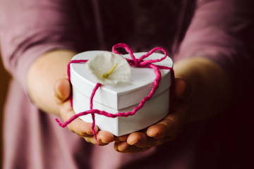 Hände halten oder überreichen eine kleine weiße Schachtel in Herzform mit einer rosa, pink Schleife und einer Blume. Ein Geschenk, geringe Schärfentiefe