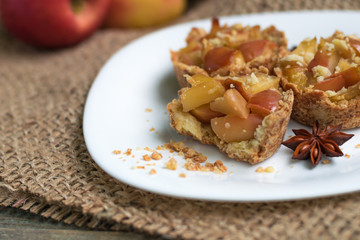 Crispy mini tartlets made from shortcrust pastry with apples, homemade sweets with anise and cinnamon. Serving on a white plate, on a wooden background. Natural eco sweets concept, warm cozy tea party