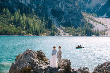 The bride and groom are standing on stones overlooking the Lago di Braies in Italy. Destination wedding in Europe, on Braies lake. Loving newlyweds walk against the backdrop of amazing nature.