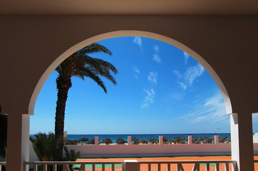 View of palm tree, deck chairs, beach and blue sky in clear weather from under the arch of the building