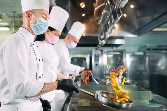 Chefs in protective masks and gloves prepare food in the kitchen of a restaurant or hotel.