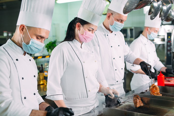 Chefs in protective masks and gloves prepare food in the kitchen of a restaurant or hotel.
