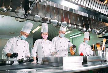 Chefs in protective masks and gloves prepare food in the kitchen of a restaurant or hotel.