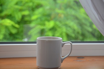 A white cup of tea or coffee stands on a wooden window sill against the background of a summer garden